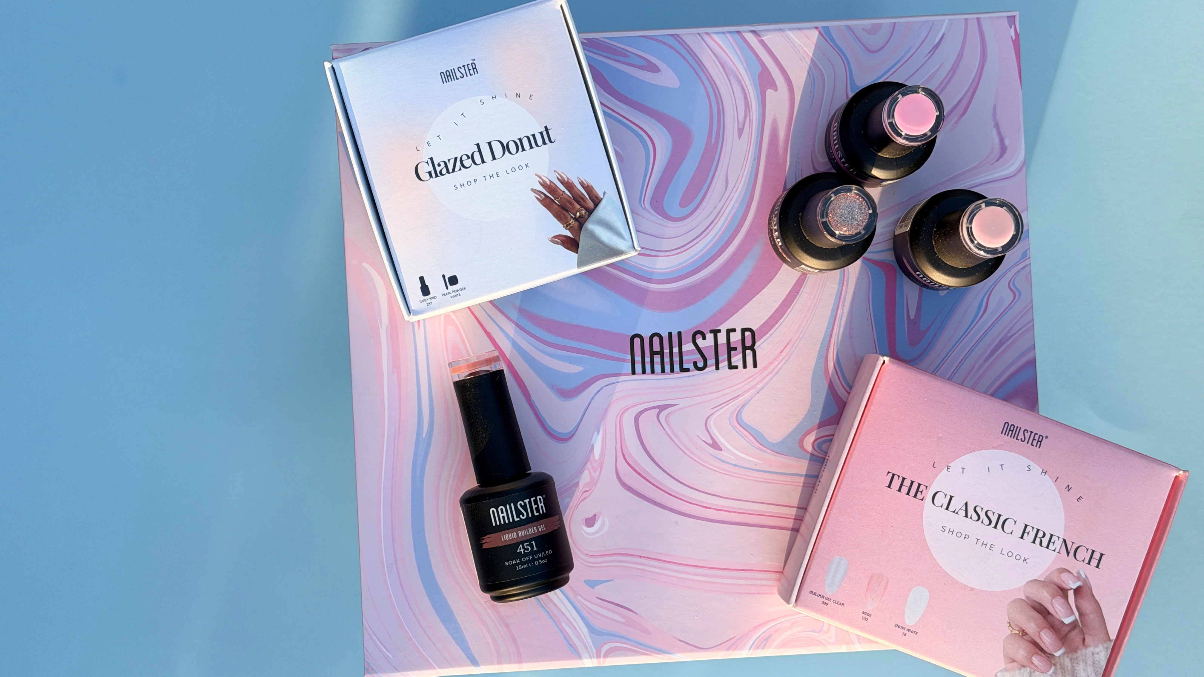 Nailster negleprodukter: Glazed Donut og Classic French sæt, builder gel, og tre neglelakflasker på en lyserød marmoreret æske.