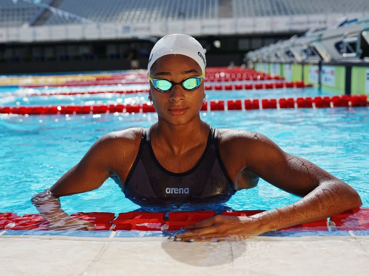 En svømmer med hvid badehætte og svømmebriller i en Arena-badedragt holder sig i kanten af en swimmingpool.