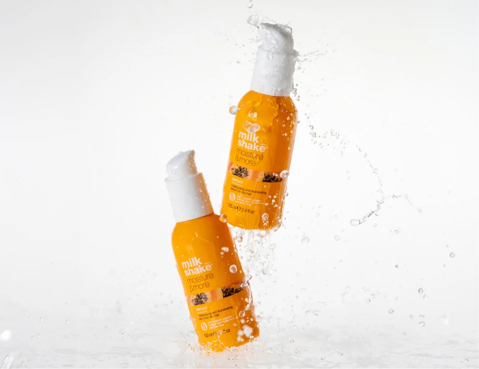 Her ses to milk_shake Moisture & More Serum i orange flasker med hvide pumper, omgivet af vandstænk mod en hvid baggrund.