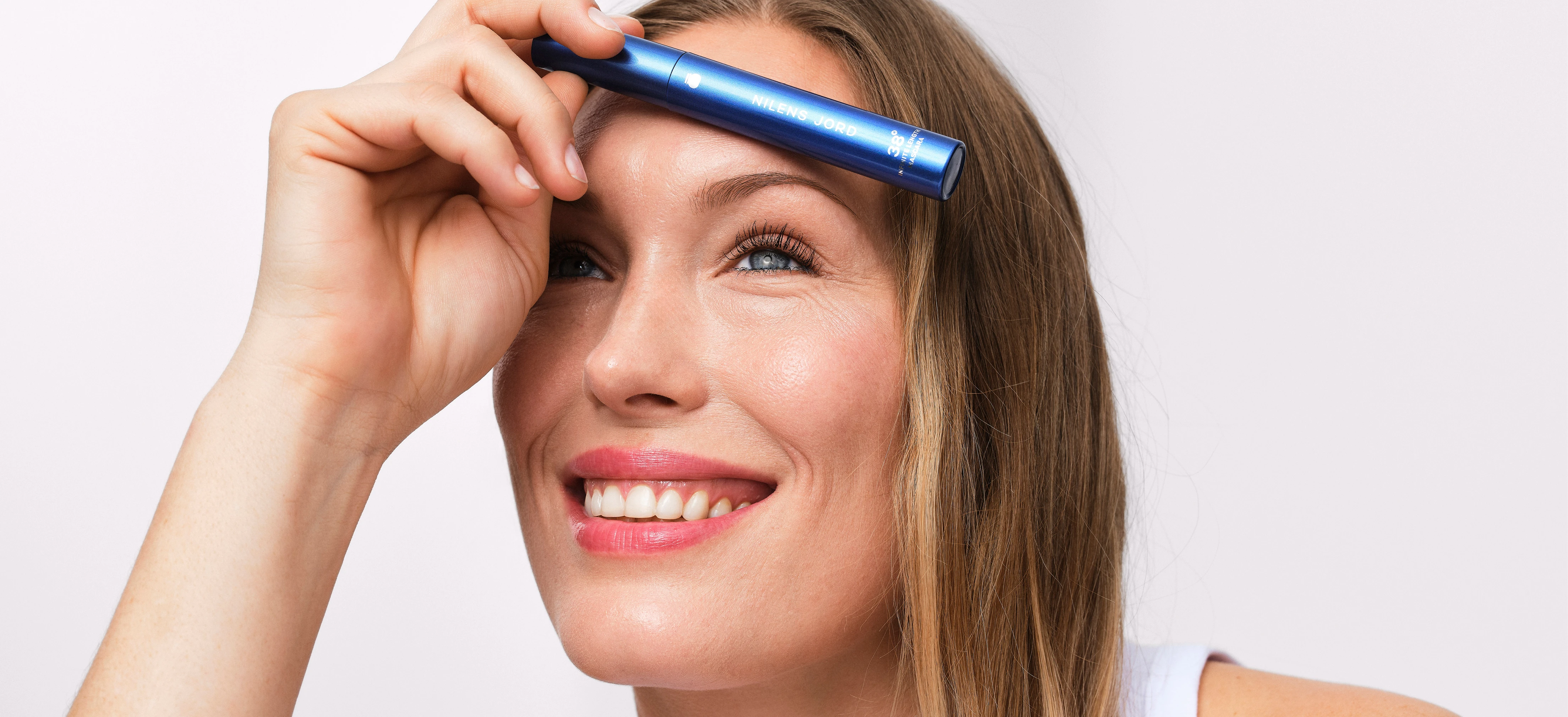 Her ses et billede af en person mod en lys baggrund, der holder en blå mascara fra Nilens Jord op foran panden. Personen smiler, og på produktet står teksten “NILENS JORD 38° Mascara”.