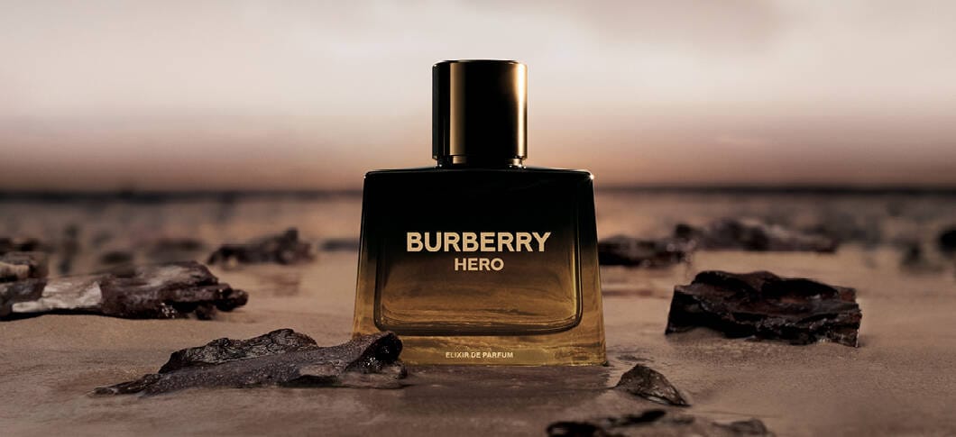 Her ses et billede af en parfumeflakon placeret på en strandlignende overflade med sten mod en mørk baggrund. På flakonen står teksten “BURBERRY HERO” samt “ELIXIR DE PARFUM”.
