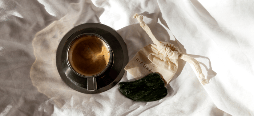 Kop med mørk te eller kaffe på en sort underkop ligger på lyst stof sammen med en grøn gua sha-sten og en lille stofpose fra A PURE MIND