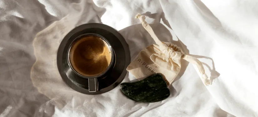 Kop med mørk te eller kaffe på en sort underkop ligger på lyst stof sammen med en grøn gua sha-sten og en lille stofpose fra A PURE MIND