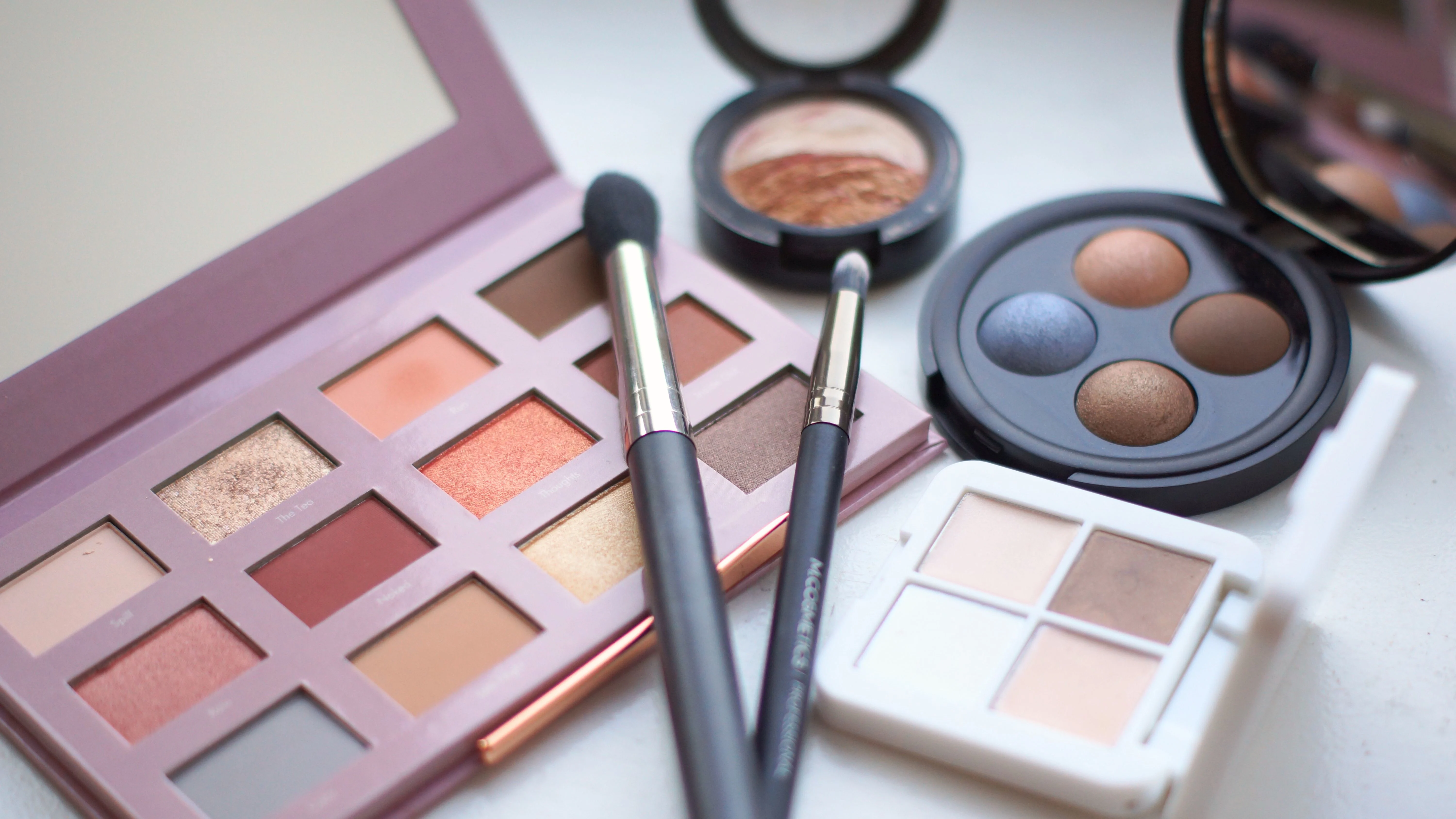Et lilla øjenskyggepalet med spejl og pensler, omgivet af andre makeup-kompakter i forskellige farver.