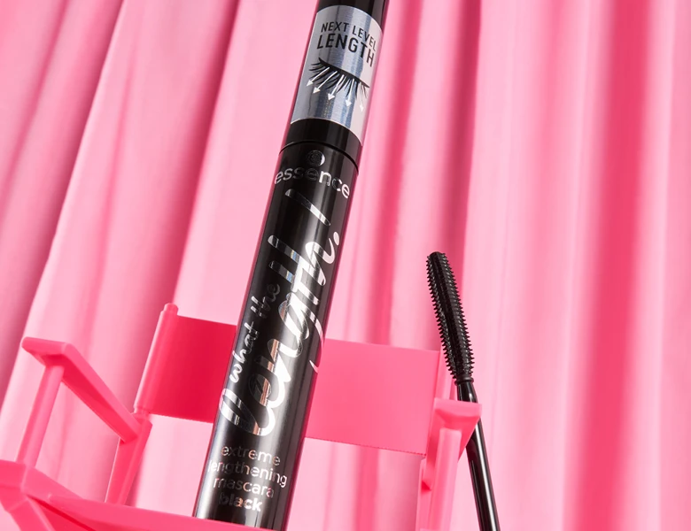Her ses et billede af en mascara fra essence placeret mod en pink baggrund med folder. På produktet står teksten “essence what the fake! extreme lengthening mascara black” og “NEXT LEVEL LENGTH”. En mascarabørste er vist ved siden af.