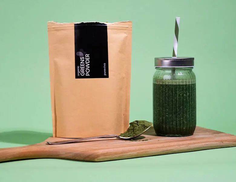 Her ses en grøn baggrund med et produkt i en brun emballage ved siden af en grøn smoothie i glad med sugerør. Det hele står på et træskærebræt