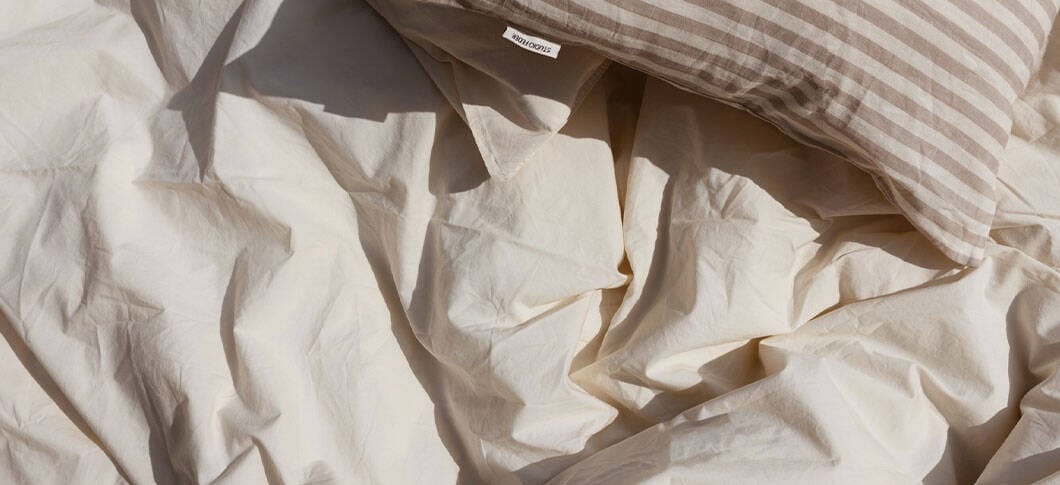 En seng med krøllet, lyst sengetøj i bløde beige og cremefarvede nuancer. Stoffet ligger i naturlige folder og giver et afslappet, uopredt udtryk. Øverst til højre ligger en pude med stribet betræk i lyse og lidt mørkere beige toner. Lyset falder blødt ind og fremhæver teksturen i stoffet, hvilket skaber en rolig og varm stemning.