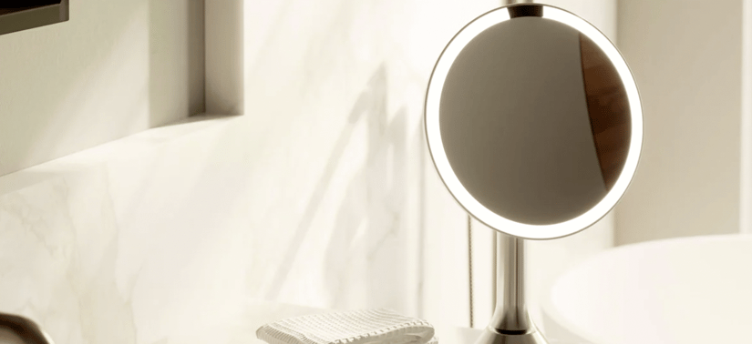 Et lyst og minimalistisk badeværelsesmiljø i beige og cremefarvede nuancer. I forgrunden står et rundt, fritstående makeup-spejl med indbygget lys langs kanten og en slank metalfod. Spejlet er tændt og danner en blød, cirkulær glød. Til højre anes kanten af en hvid håndvask. Foran spejlet ligger en lille, lys klud eller vaskesvamp med rillet tekstur. Baggrunden består af glatte vægge og rene linjer, som giver et roligt og moderne udtryk.