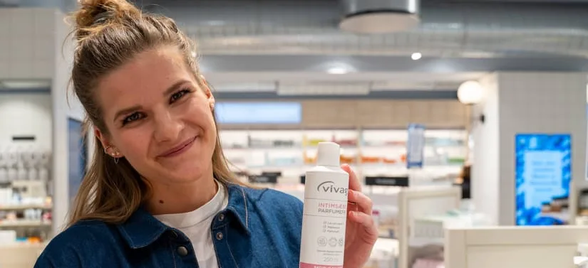 Smilende kvinde i en butik holder en hvid flaske med intimplejeprodukt op mod kameraet. Hun står foran hylder med produkter i et lyst butiksmiljø.