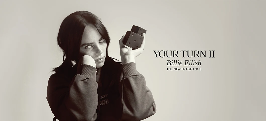 Her ses et billede med en person til venstre, der holder en parfumeflakon, mod en lys baggrund. Til højre står teksten “YOUR TURN II Billie Eilish” samt “THE NEW FRAGRANCE”.