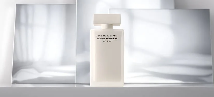 Her ses et billede af en parfumeflakon placeret centralt mod en lys, neutral baggrund med spejlende flader. På flakonen står teksten “PURE MUSC BLANC narciso rodriguez for her”. Flakonen er hvid med et enkelt, firkantet design og låg i samme farve.