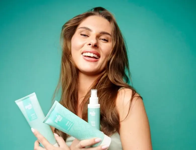 Her ses en kvinde, der holder Yuaia Hair produkter i grøn emballage på en turkis baggrund.