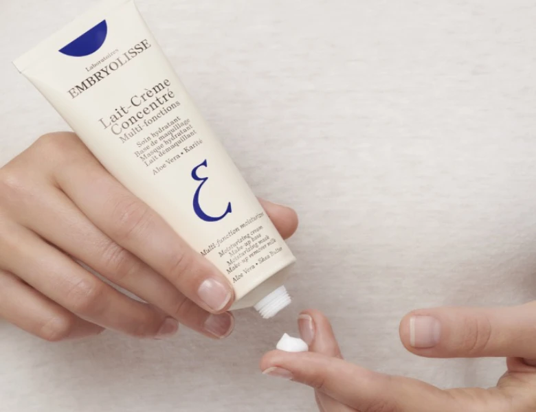 Her ses en hånd, der holder en tube Embryolisse Lait-Crème Concentré, mens creme påføres en finger, mod en lys baggrund.