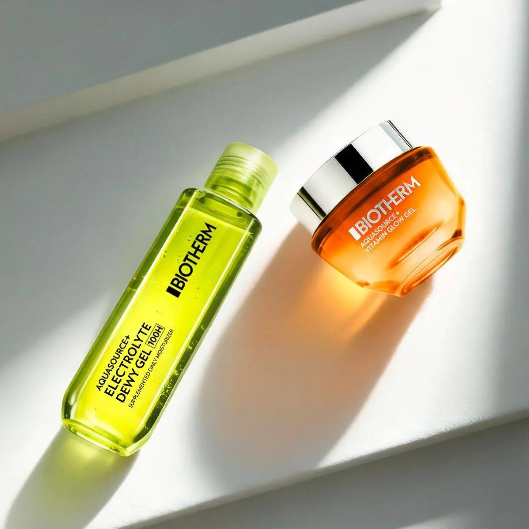 To Biotherm-hudplejeprodukter ligger på en lys overflade oplyst af sollys. Til venstre ses en grøn flaske med Aquasource+ Electrolyte Dewy Gel, og til højre en orange krukke med Aquasource Vitamin Glow Gel med sølvfarvet låg. Produkterne kaster bløde skygger og har et friskt og energifyldt udtryk med fokus på fugt og glød til huden.