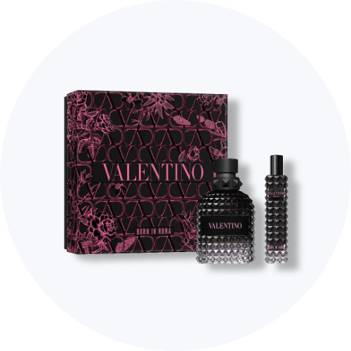 Her ses et billede med en gaveæske fra Valentino placeret mod en lys, rund baggrund. Ved siden af æsken står to parfumeflakoner med teksten “VALENTINO BORN IN ROMA”. Produkterne er i sorte nuancer med nitteinspireret design og pink detaljer.