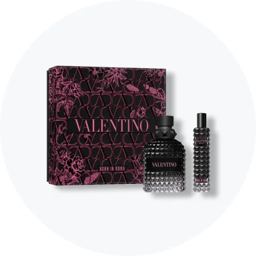 Her ses et billede med en gaveæske fra Valentino placeret mod en lys, rund baggrund. Ved siden af æsken står to parfumeflakoner med teksten “VALENTINO BORN IN ROMA”. Produkterne er i sorte nuancer med nitteinspireret design og pink detaljer.