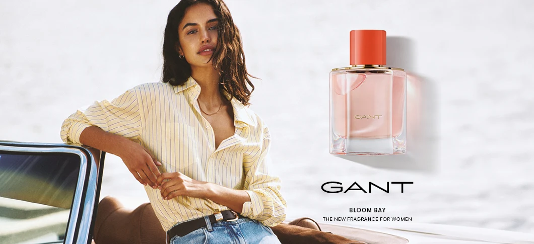 Her ses et billede med en person til venstre, der læner sig op ad en bil mod en lys baggrund, samt en parfumeflakon til højre. På billedet står teksten “GANT”, “BLOOM BAY” og “THE NEW FRAGRANCE FOR WOMEN”. Flakonen har teksten “GANT” på fronten.