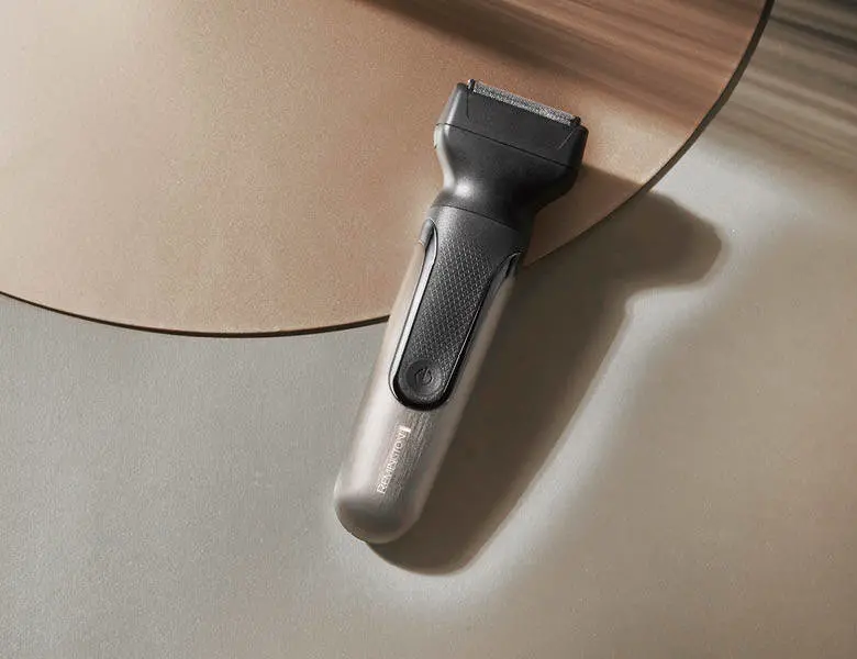 Her ses et billede af en elektrisk trimmer placeret på en lys overflade med en rund plade i baggrunden. Trimmeren er i grå og sort design med en tænd/sluk-knap på fronten.