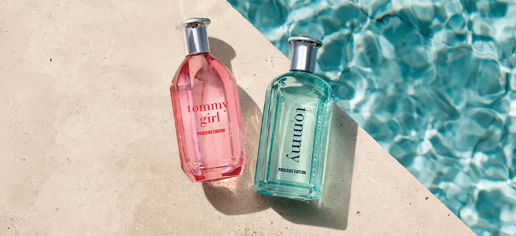 Her ses et billede med to parfumeflakoner placeret på en lys overflade ved siden af vand. På den ene flakon står “tommy girl poolside edition”, og på den anden står “tommy poolside edition”. Flakonerne er i pink og turkis nuance med sølvfarvede låg.
