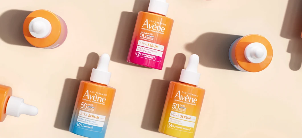Her ses Avène Ultra Serum produkter i orange, pink og blå emballage på en lys beige baggrund.