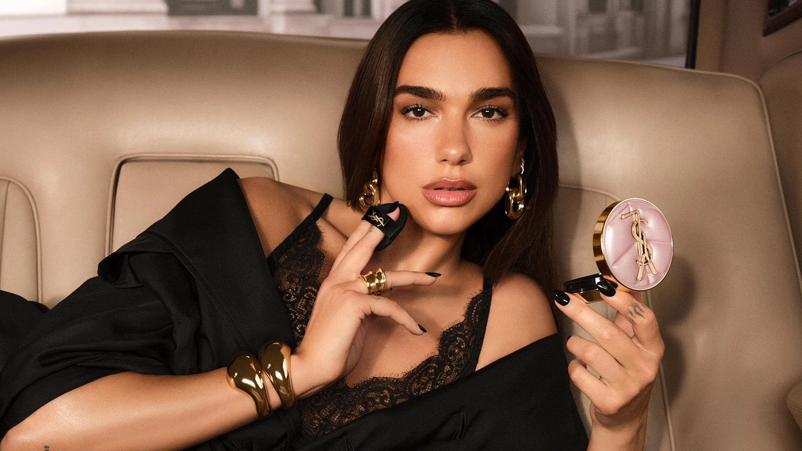 Dua Lipa holder et lyserødt YSL-pudder og en sort YSL-applikator, iført en sort blondebluse og guldsmykker.