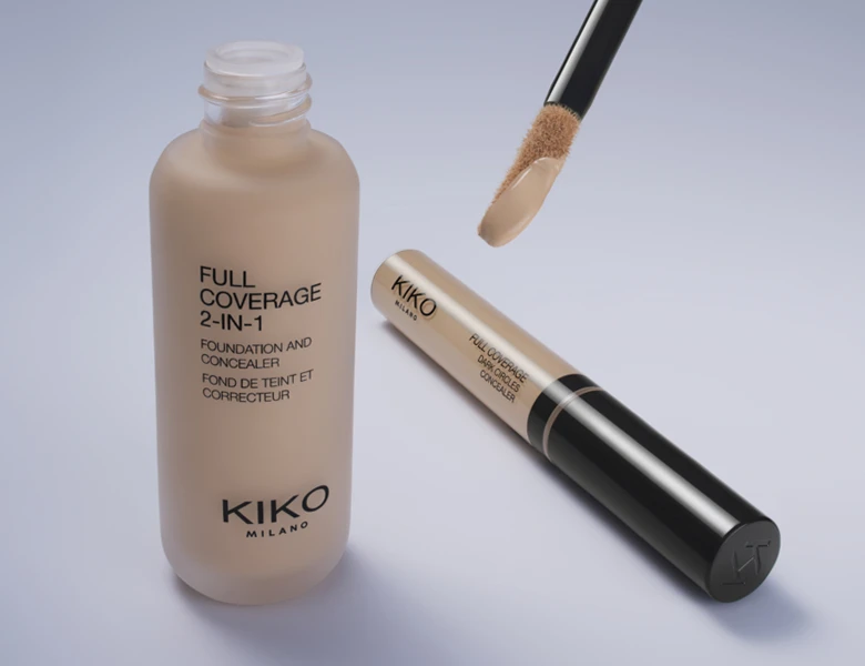 Her ses et billede med makeup-produkter fra KIKO Milano placeret mod en lys baggrund. Der ses en foundation og en concealer med applikator, og på produkterne står teksten “FULL COVERAGE 2-IN-1 Foundation and Concealer”.