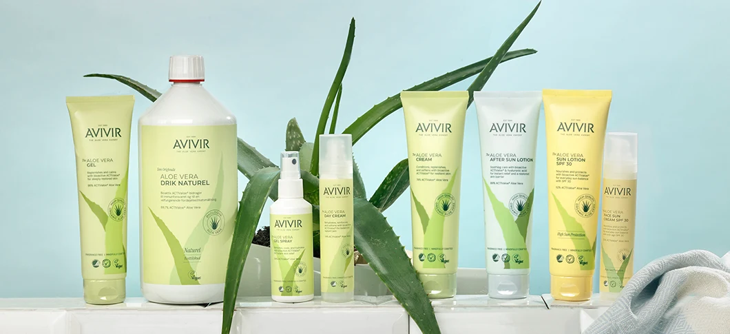 Her ses et udvalg af AVIVIR Aloe Vera-produkter i grønne, hvide og gule emballager omgivet af aloe vera-planter mod en lyseblå baggrund.