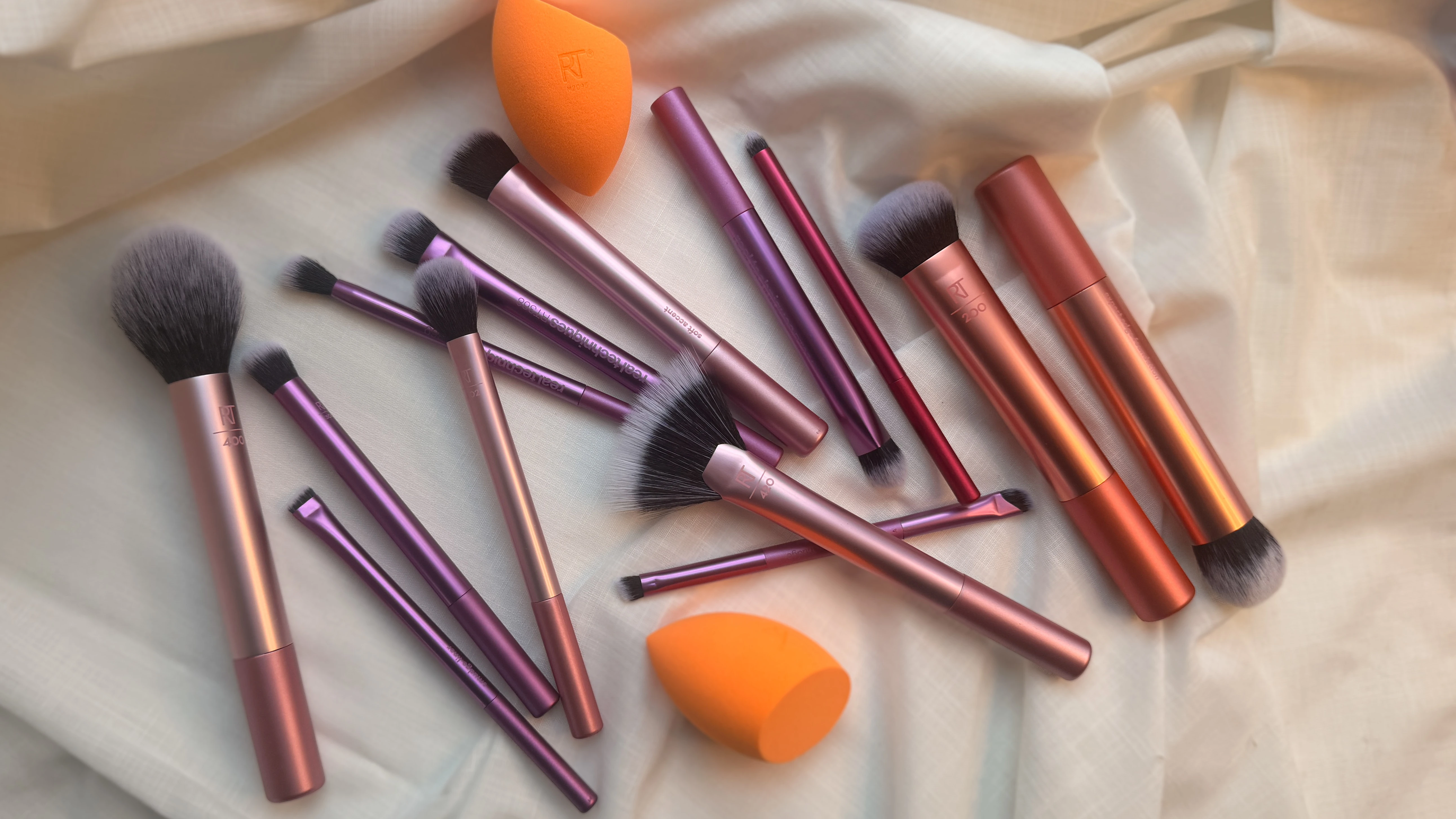 Makeupbørster og makeup-svampe fra Real Techniques med rosaguld og lilla håndtag, spredt ud på lyst stof.