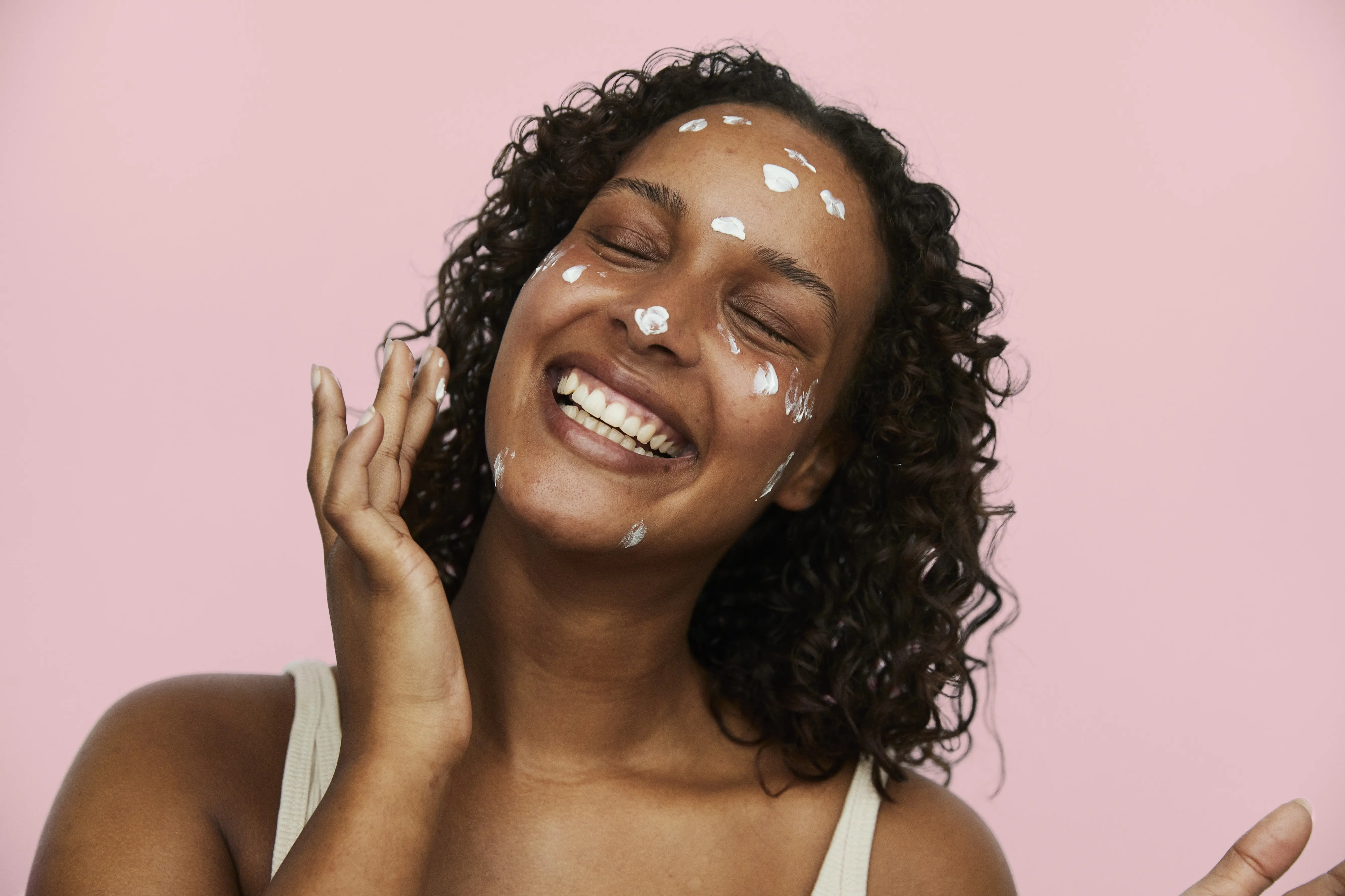 Her ses en smilende person med mørkt, krøllet hår og lukkede øjne. Personen har hvid creme påført flere steder i ansigtet og holder den ene hånd ved kinden mod en rosa baggrund.