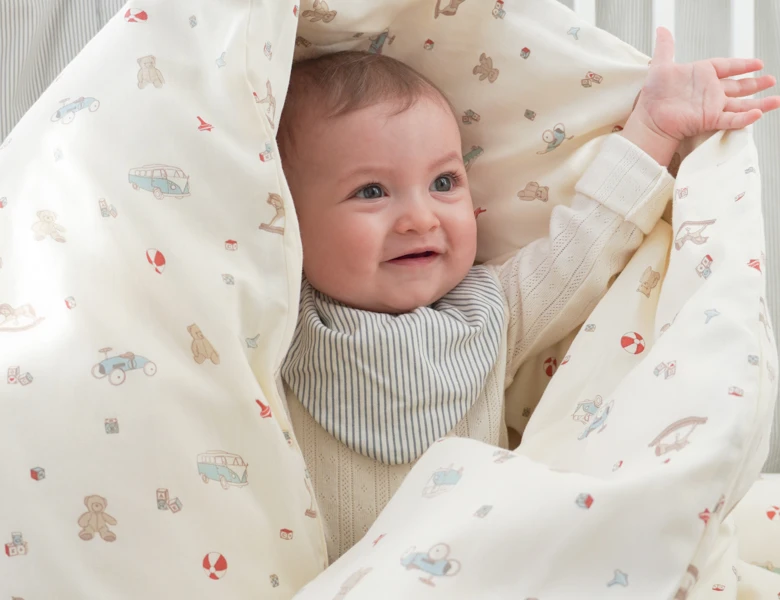 Smilende baby ligger svøbt i et lyst tæppe med små illustrationer og løfter den ene arm.