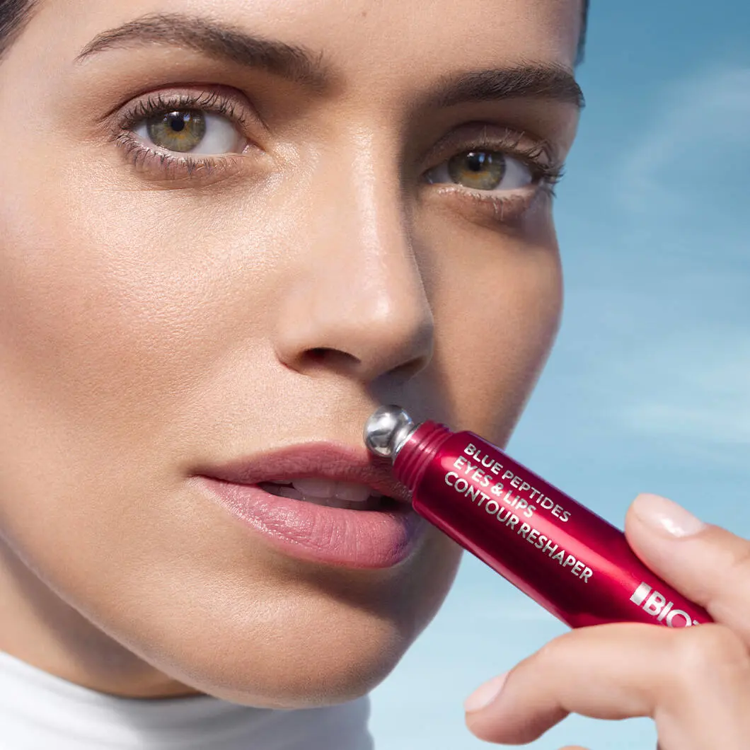 Nærbillede af en kvindes ansigt med fokus på øjne og læber. Hun holder et rødt kosmetikprodukt med metalrulle op mod overlæben. På produktet står der “Blue Peptides Eyes & Lips Contour Reshaper”. Kvinden har naturlig makeup og ser direkte mod kameraet. Baggrunden er lyseblå og giver et rent, friskt udtryk.