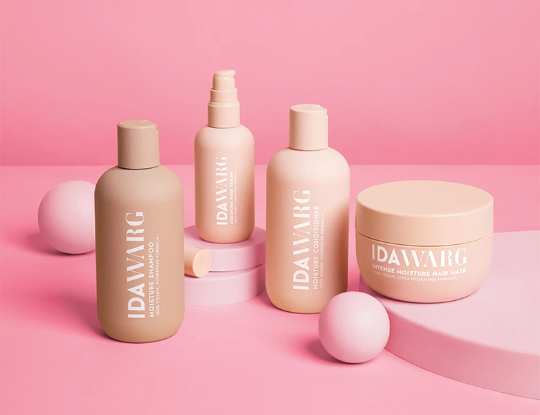 Her ses IDA WARG hårplejeprodukter – shampoo, conditioner og hårmaske i beige emballage på en lyserød baggrund.