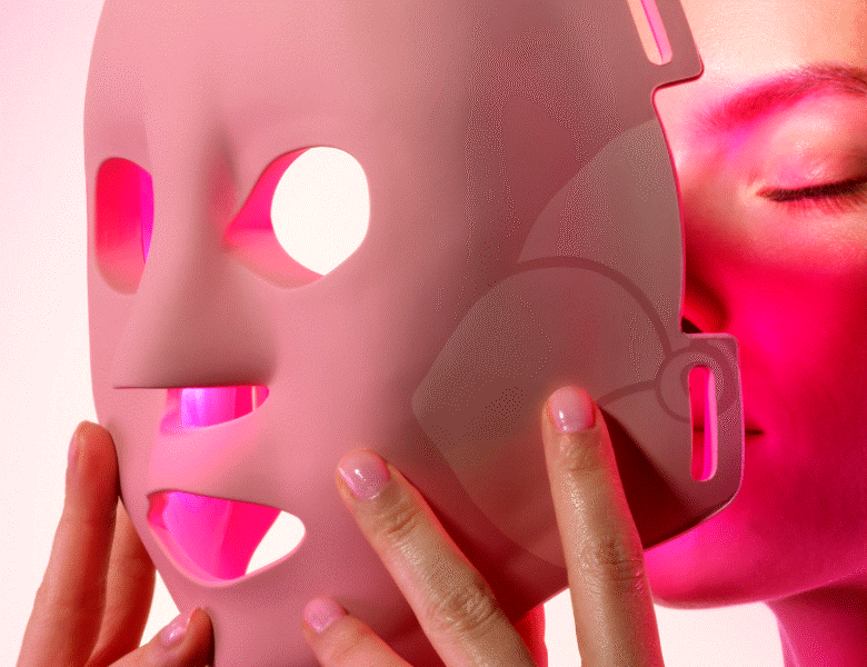 Her ses et close-up billede af en kvinde, som er ved at tage en LED-ansigtsmaske på. Masken er lyserød og har rødt lys.