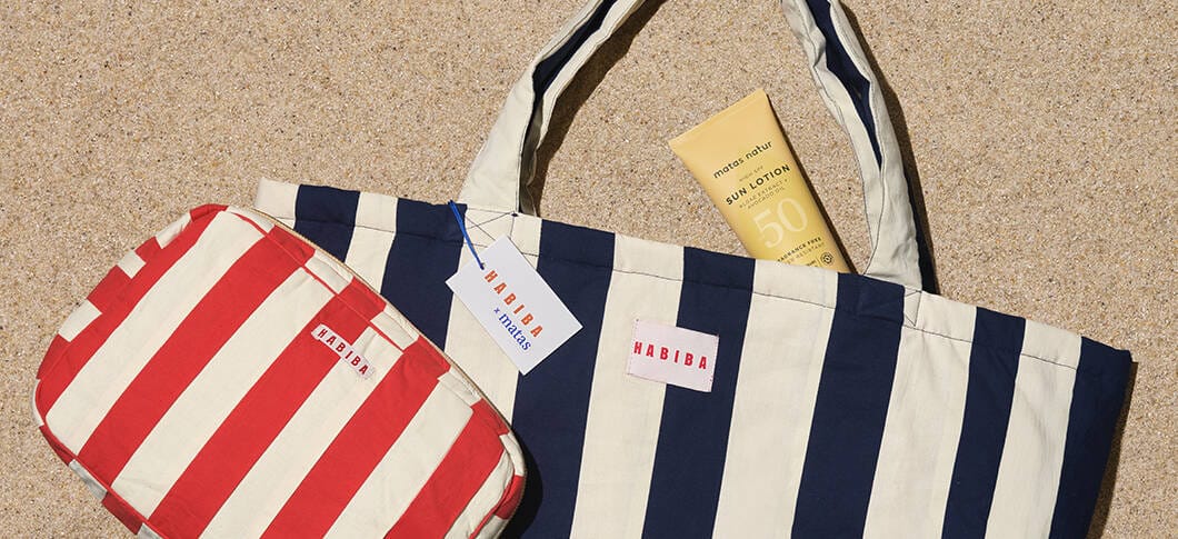 En strandscene set oppefra med sand som baggrund. I midten ligger en stor stofpose med brede striber i mørkeblå og cremefarve og teksten “HABIBA” på en lille label. I posen stikker en gul solcreme med teksten “Sun Lotion SPF 50” op. Til venstre ligger en mindre toilettaske i samme design, men med røde og cremefarvede striber, også med “HABIBA”-label. På tasken hænger et prismærke med teksten “HABIBA x matas”. Farverne er sommerlige og maritime.