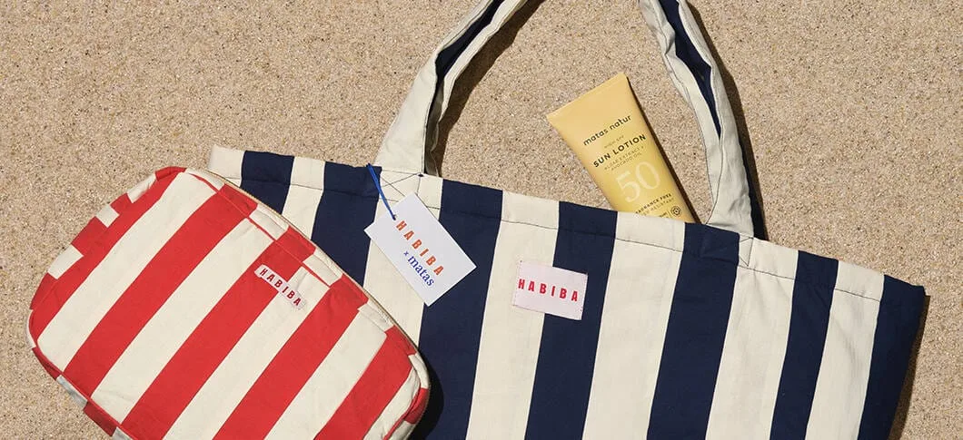En strandscene set oppefra med sand som baggrund. I midten ligger en stor stofpose med brede striber i mørkeblå og cremefarve og teksten “HABIBA” på en lille label. I posen stikker en gul solcreme med teksten “Sun Lotion SPF 50” op. Til venstre ligger en mindre toilettaske i samme design, men med røde og cremefarvede striber, også med “HABIBA”-label. På tasken hænger et prismærke med teksten “HABIBA x matas”. Farverne er sommerlige og maritime.