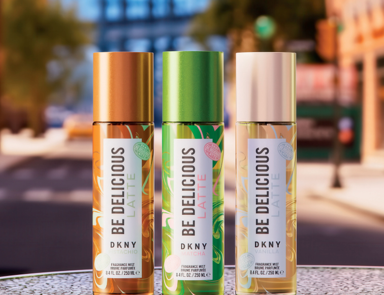 Her ses et billede med tre høje parfumemists fra DKNY placeret side om side mod en sløret bybaggrund. På produkterne står teksten “BE DELICIOUS LATTE DKNY”, og varianterne er i brune, grønne og lyse nuancer med matchende låg.