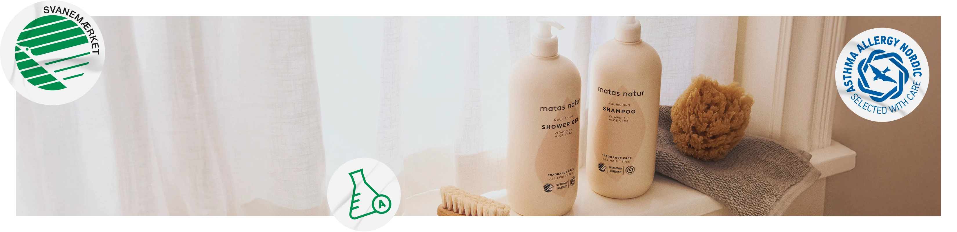 Her ses Matas Natur Shower Gel og Shampoo på en vindueskarm foran lyse gardiner. Ved siden af produkterne ligger en træbørste, et gråt håndklæde og en natursvamp. Billedet viser også Svanemærket, Asthma Allergy Nordic og A-kolben fra Forbrugerrådet Tænk.