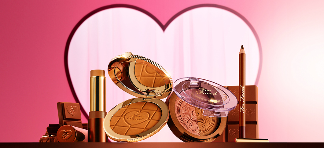 Her ses et billede med flere makeup-produkter fra Too Faced placeret mod en pink baggrund med et hjerteformet motiv. Produkterne inkluderer blandt andet bronzer, blush, læbeprodukt og eyeliner i gyldne og brune nuancer. På produkterne står teksten “Too Faced”.