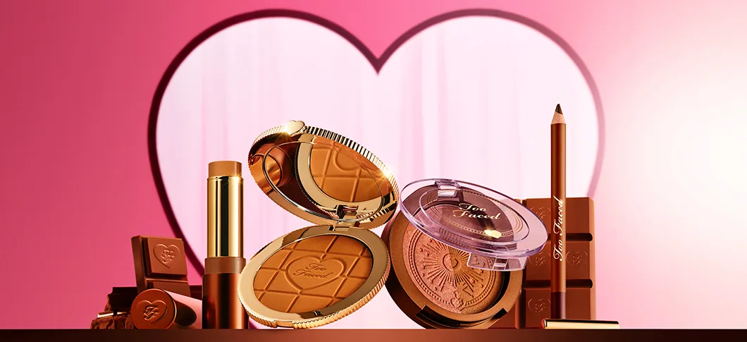 Her ses et billede med flere makeup-produkter fra Too Faced placeret mod en pink baggrund med et hjerteformet motiv. Produkterne inkluderer blandt andet bronzer, blush, læbeprodukt og eyeliner i gyldne og brune nuancer. På produkterne står teksten “Too Faced”.