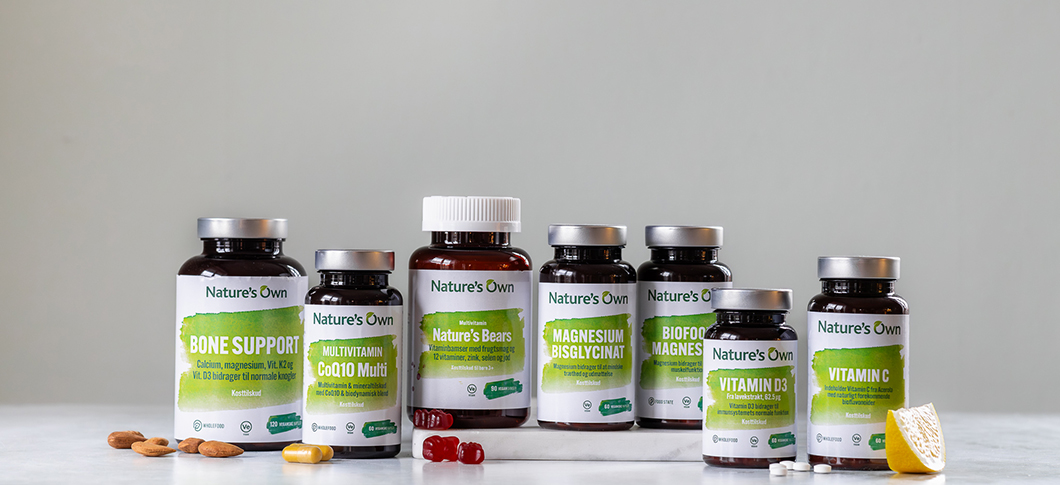 Flere Nature’s Own kosttilskud står samlet på en lys overflade foran en neutral grå baggrund, blandt andet Bone Support, CoQ10, vitaminbjørne, magnesium, biofood magnesium, vitamin D3 og vitamin C, med mandler, vingummier, tabletter og en citron foran