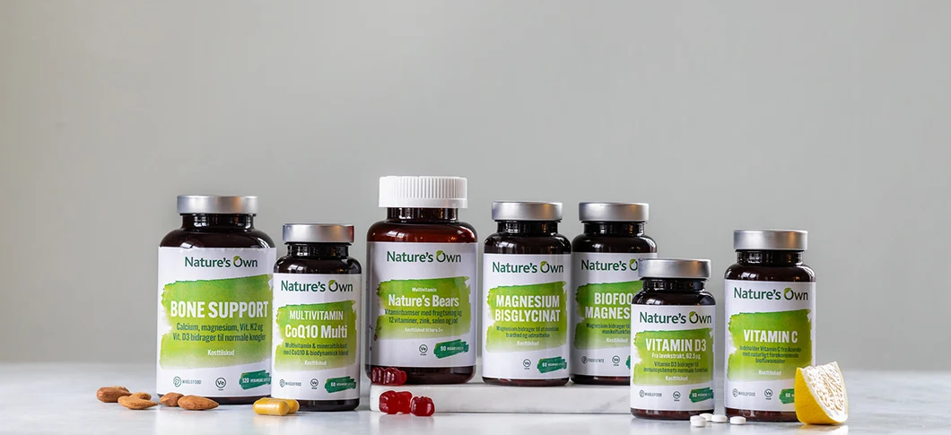Flere Nature’s Own kosttilskud står samlet på en lys overflade foran en neutral grå baggrund, blandt andet Bone Support, CoQ10, vitaminbjørne, magnesium, biofood magnesium, vitamin D3 og vitamin C, med mandler, vingummier, tabletter og en citron foran