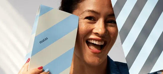 Her ses et billede af en person, der smiler og holder to æsker med Matas’ karakteristiske stribede design i blå, beige og grå nuancer op foran ansigtet. På den ene æske står teksten “matas”.