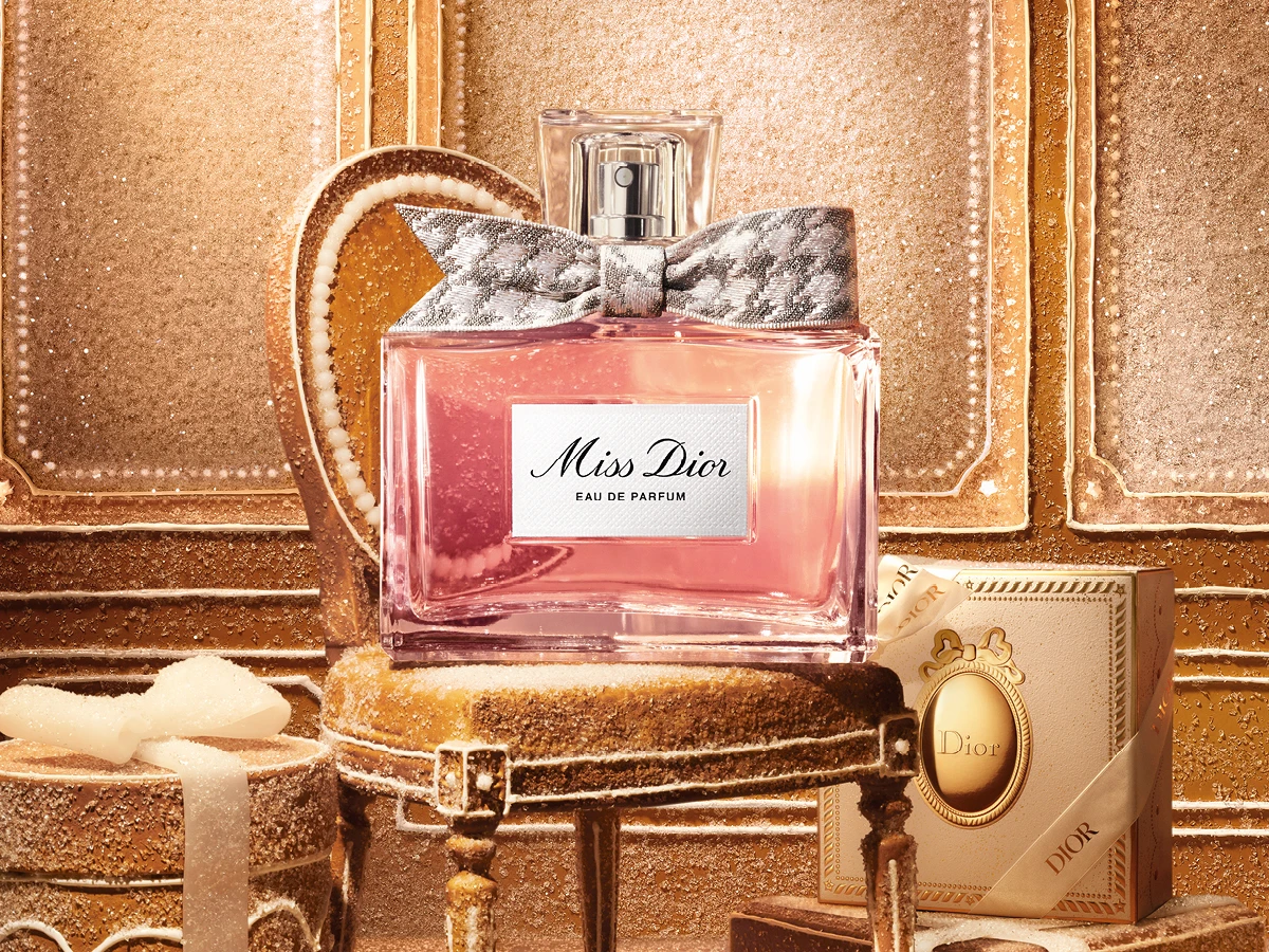 Miss Dior Eau de Parfum-flaske med mønstret sløjfe, placeret på gylden glitrende stol omgivet af Dior julepynt.