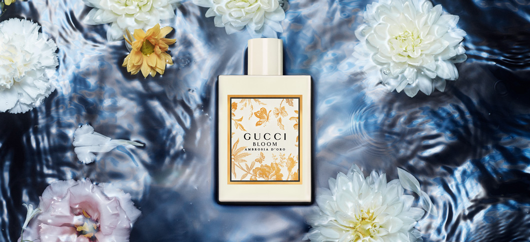 Her ses et billede af en parfumeflakon med teksten “GUCCI BLOOM AMBROSIA DI FIORI” placeret centralt i vand med blomster omkring. Flakonen er i en lys, mat farve med et dekorativt label.