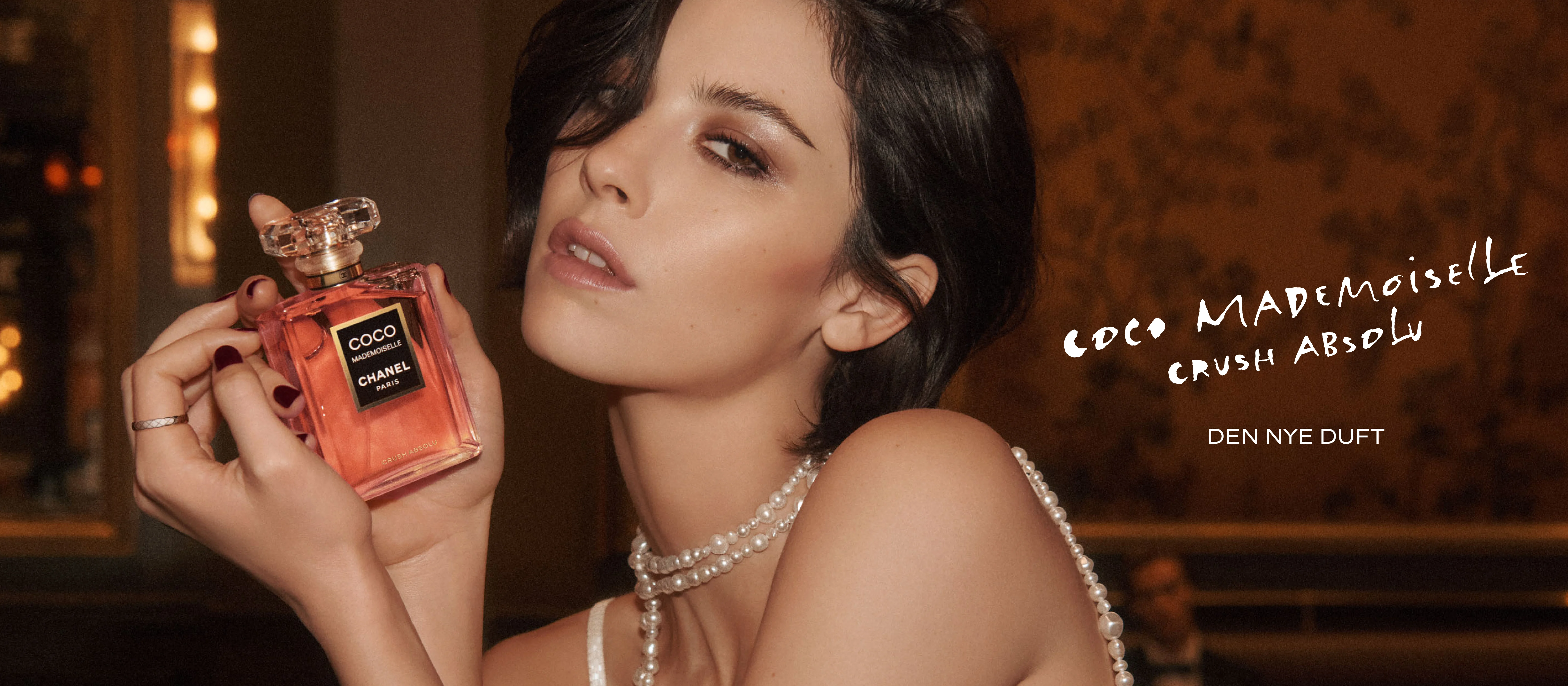 Her ses et kampagnebillede for Chanel Coco Mademoiselle Crush Absolu i et varmt og eksklusivt univers. En person holder den rosa parfumeflakon tæt ved ansigtet og er stylet med en lys kjole og perlesmykker. Øverst står CHANEL, mens teksten “Coco Mademoiselle Crush Absolu” og “Den nye duft” fremhæver lanceringen.