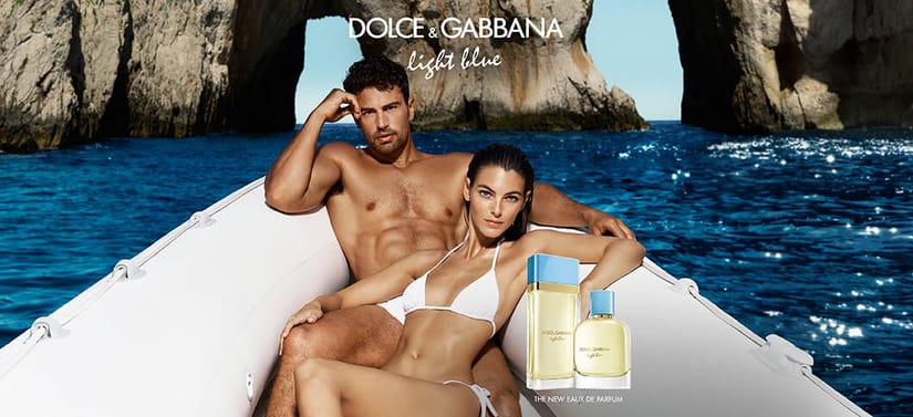 Mand og kvinde i badetøj sidder tæt sammen i en båd på blåt hav foran klipper, med Dolce & Gabbana Light Blue parfumer vist i forgrunden.