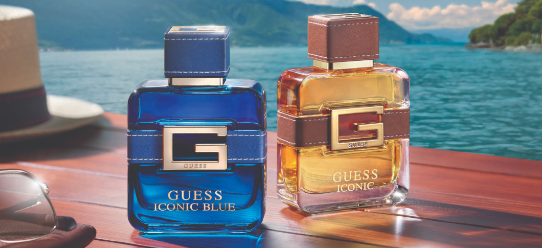 Her ses et billede med to parfumeflakoner fra Guess placeret på et træbord ved vandet. På flakonerne står teksten “GUESS ICONIC BLUE” og “GUESS ICONIC”. Flakonerne er i blå og gyldne nuancer med store “G”-detaljer på fronten.