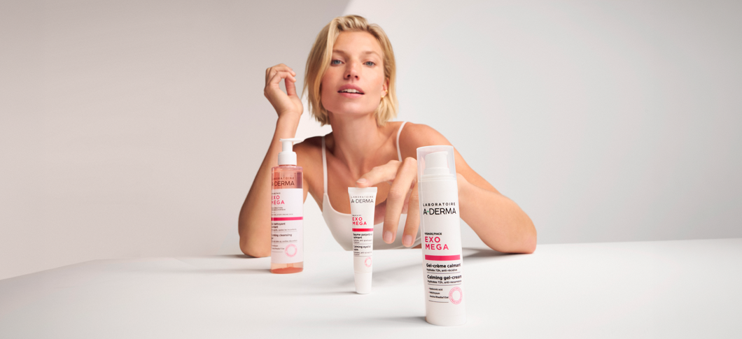 Her ses en kvinde sammen med A-DERMA Exomega hudplejeprodukter i hvid emballage på en lys baggrund.