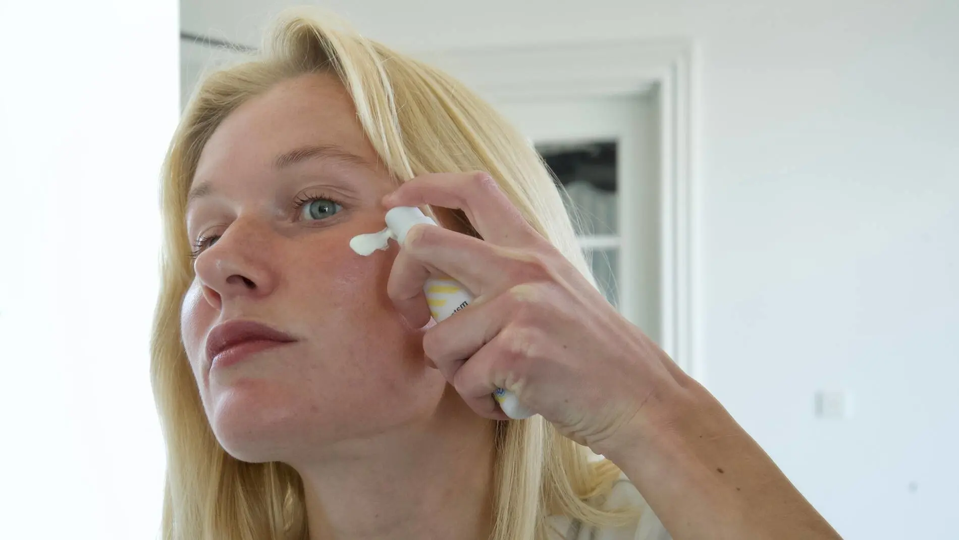 En person med lyst hår påfører ansigtscreme på kinden med fingrene, mens personen holder en flaske med solcreme fra Matas Striber i den anden hånd. Personen står indendørs foran et spejl i et lyst badeværelse og ser fokuseret mod spejlet. Solcremen har hvid emballage med gule striber og flere certificeringsikoner nederst på flasken.
