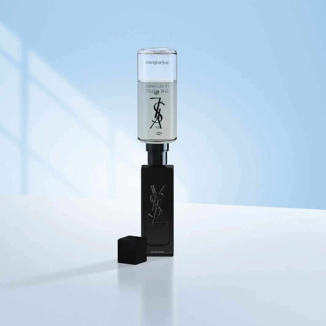 En sort Yves Saint Laurent MYSLF-parfumeflaske står på en lys overflade foran en blød lyseblå baggrund. Oven på flasken er en gennemsigtig refillbeholder monteret for at vise genopfyldning af parfumen. Flaskens sorte låg ligger ved siden af. Billedet har et stilrent og eksklusivt udtryk med fokus på luksusparfume og refill-løsninger.